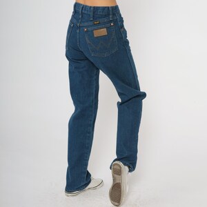 May include: Dark blue denim Wrangler jeans with a brown leather patch on the back pocket. The jeans have a classic straight leg fit and are worn with white sneakers.