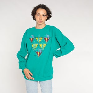 May include: Teal-colored sweatshirt with a triangular design featuring palm trees and cacti. The graphic includes yellow, purple, and red accents. The long-sleeved top is a casual style.