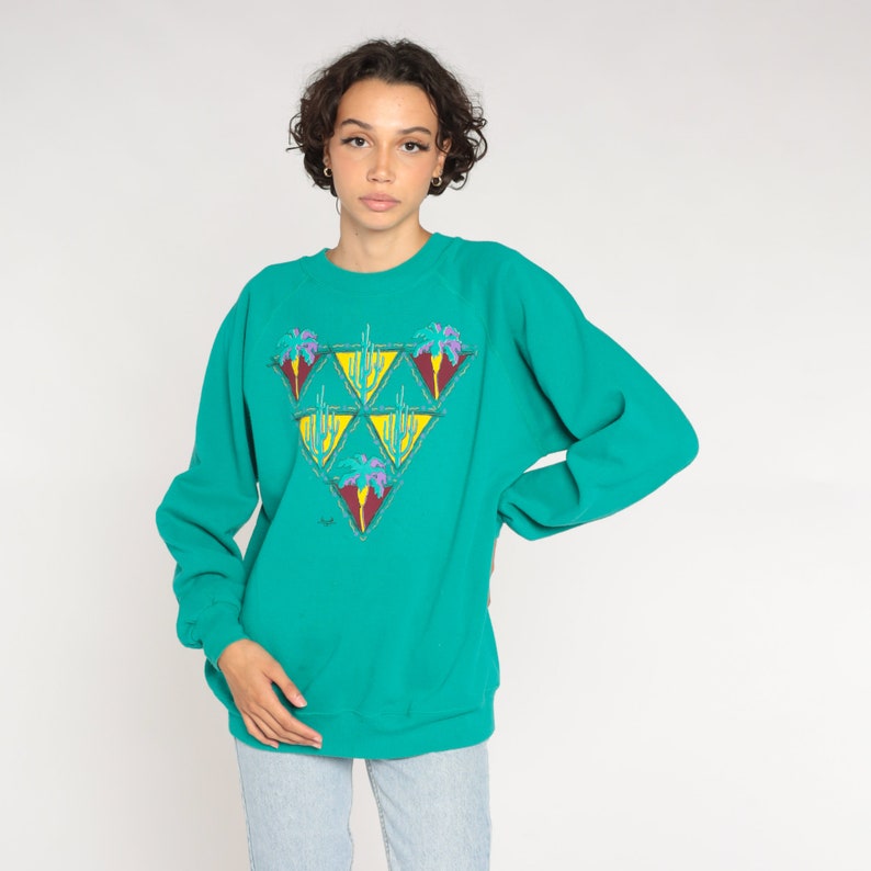 May include: Teal-colored sweatshirt with a triangular design featuring palm trees and cacti. The graphic includes yellow, purple, and red accents. The long-sleeved top is a casual style.