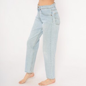 May include: Light blue denim jeans with a high waist and straight leg. The jeans have classic five-pocket styling and a button and zipper closure. The denim is a light wash, and the jeans appear to be vintage.