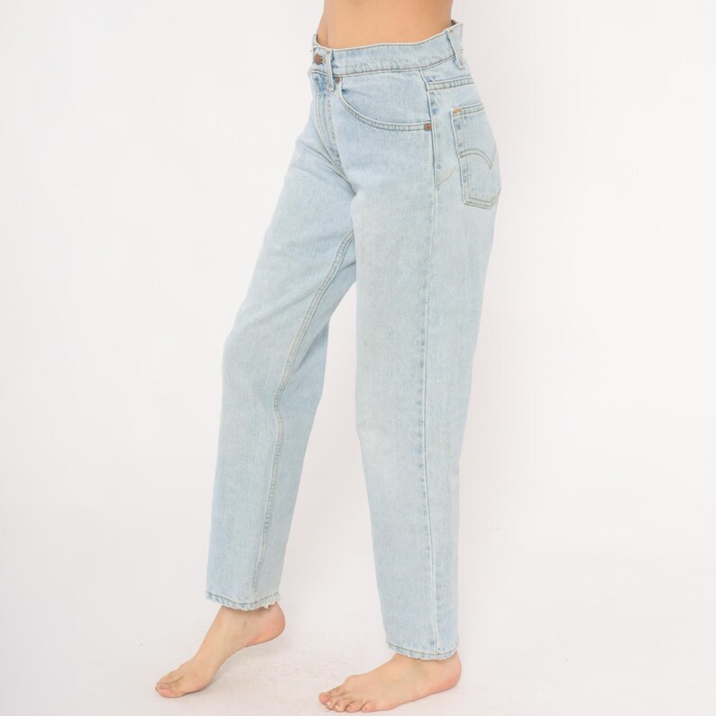 May include: Light blue denim jeans with a high waist and straight leg. The jeans have classic five-pocket styling and a button and zipper closure. The denim is a light wash, and the jeans appear to be vintage.