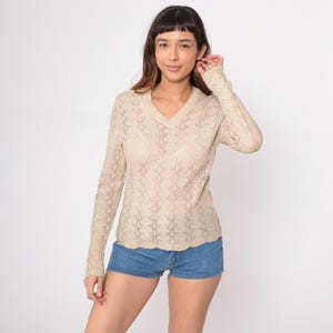 Puede incluir: Un suéter beige de manga larga con cuello en V y un delicado diseño calado. El suéter se combina con pantalones cortos de mezclilla. El look general es casual y cómodo.