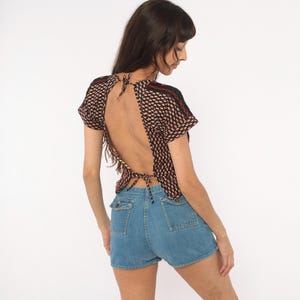 May include: A black short-sleeved top with a repeating small red and white geometric pattern, featuring an open back design with thin tie closures at the neck and waist, paired with blue denim shorts that have two front pockets with button flaps.