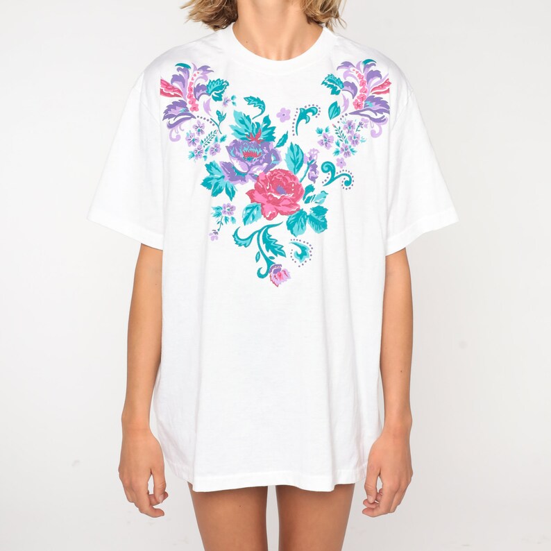 May include: White t-shirt with a floral design in shades of pink, purple, and teal.