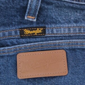 May include: Close-up of blue denim jeans featuring a back pocket with a brown leather patch and the brand name "Wrangler" embossed. The pocket has a brass button and the Wrangler logo is also on a small black label.