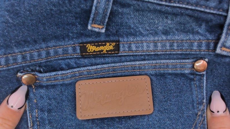 May include: Close-up of blue denim jeans featuring a back pocket with a brown leather patch and the brand name "Wrangler" embossed. The pocket has a brass button and the Wrangler logo is also on a small black label.