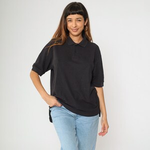 Pode incluir: Uma camisa polo preta com mangas curtas e gola. A camisa é feita de um material macio e é usada com jeans azul claro. A modelo está sorrindo e olhando para a câmera.