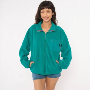 May include: A teal fleece jacket featuring a full-length front zipper, a high collar with thin pink and white trim, long sleeves, and a single zippered chest pocket on the left side. The jacket has two side pockets and a relaxed fit design.