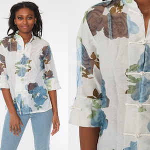 May include: White button-up shirt with a floral pattern featuring blue and brown roses. The shirt has a mandarin collar and three-quarter sleeves. It is fastened with fabric loops and buttons.