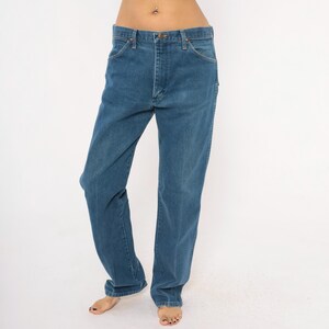 May include: Blue denim jeans with a classic five-pocket design. The jeans have a straight leg cut and a button and zipper closure. The denim is a medium wash, and the jeans appear to be vintage.