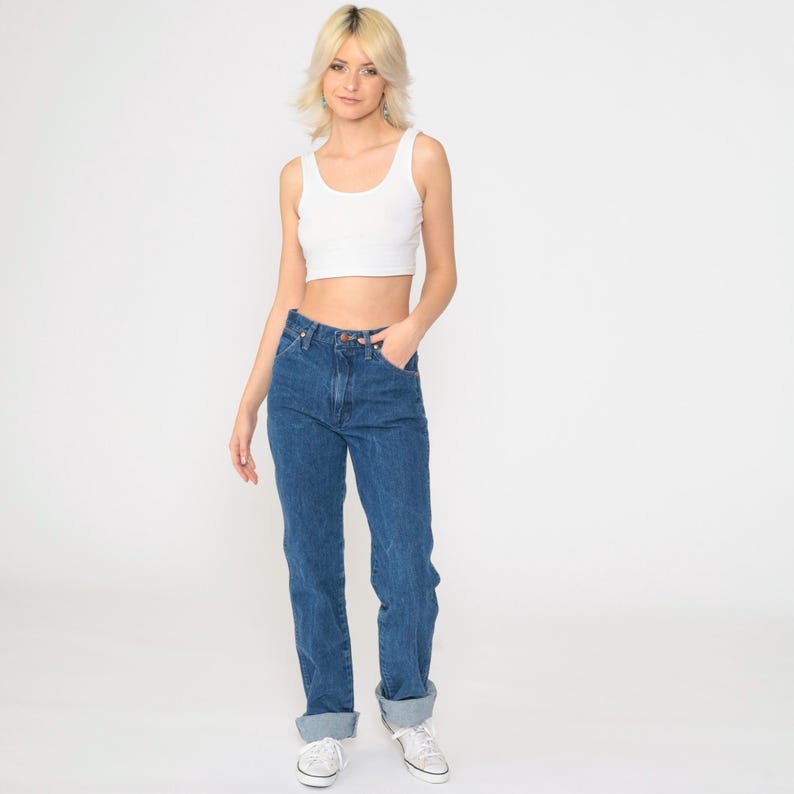 May include: A woman wearing a white tank top and blue denim jeans with the cuffs rolled up. She is standing with her hand in her pocket and looking at the camera.