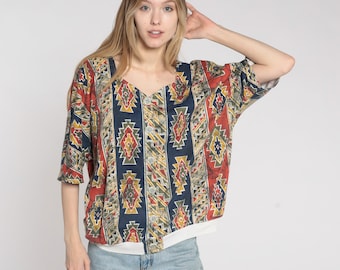May include: A button-down shirt with a V-neck and short sleeves. The shirt features a colorful geometric pattern in red, navy blue, yellow, and green. The shirt is worn over a white undershirt and paired with light blue jeans.