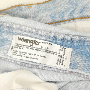May include: Close-up of light blue Wrangler jeans. The label reads "Wrangler 34 x 30". The jeans are made of 100% cotton and are exclusive of decoration. The jeans have a button and stitching.
