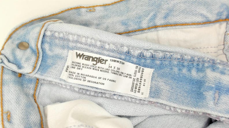 May include: Close-up of light blue Wrangler jeans. The label reads "Wrangler 34 x 30". The jeans are made of 100% cotton and are exclusive of decoration. The jeans have a button and stitching.