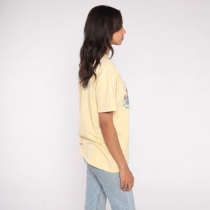 May include: A light yellow t-shirt with a graphic design. The graphic features a colorful illustration. The shirt is paired with light blue jeans. The model is standing in front of a white background.