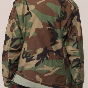May include: A long-sleeve field jacket featuring a woodland camouflage pattern in shades of olive green, brown, and black. The jacket has a fold-over collar, shoulder epaulets, and a straight hemline that reveals a light green interior lining.