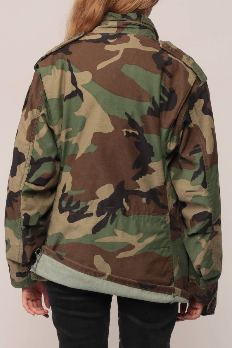 May include: A long-sleeve field jacket featuring a woodland camouflage pattern in shades of olive green, brown, and black. The jacket has a fold-over collar, shoulder epaulets, and a straight hemline that reveals a light green interior lining.