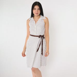 May include: A white dress with black polka dots. The sleeveless dress has a collared neckline, a brown belt tied at the waist, and a zip closure. The dress is knee-length and has a flared skirt.