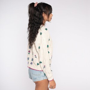 May include: A white knitted sweater with a cable knit pattern and embroidered flowers in shades of green, blue, and pink. The sweater has a crew neck and long sleeves.