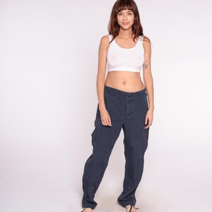 May include: A woman wearing a white tank top and navy blue cargo pants. The pants have multiple pockets and a relaxed fit. The woman is standing in front of a white background.