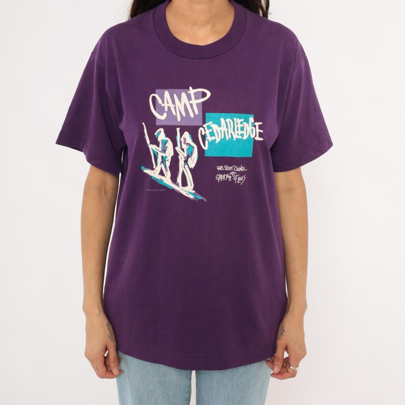 May include: A purple t-shirt with a crew neck featuring a graphic design. The design includes the words "CAMP CEDARLEGE" in white and a drawing of two figures on a surfboard. Additional text is present below the main design.