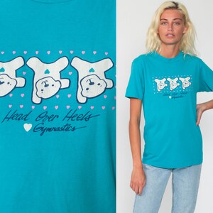May include: A turquoise blue t-shirt with a graphic of three white cartoon bears doing a handstand. The text "Head Over Heels Gymnastics" is printed below the bears.