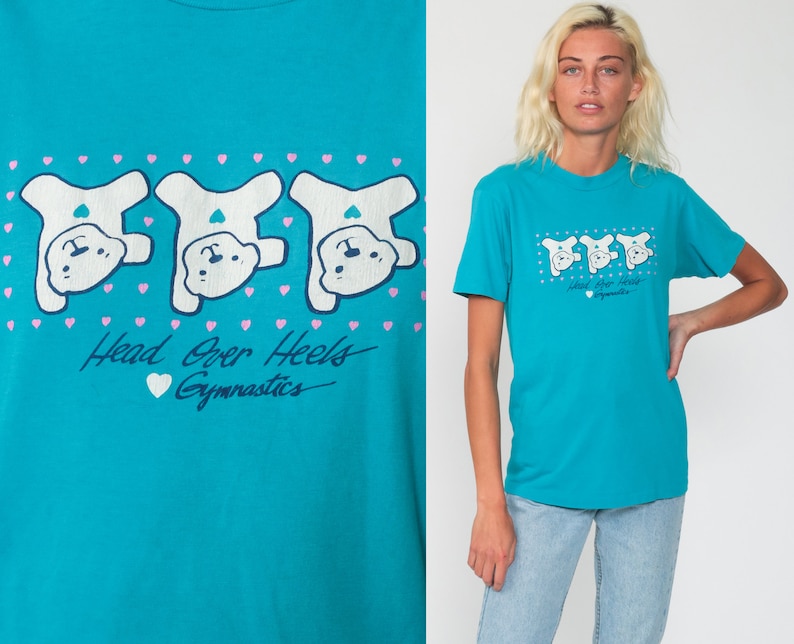 May include: A turquoise blue t-shirt with a graphic of three white cartoon bears doing a handstand. The text "Head Over Heels Gymnastics" is printed below the bears.
