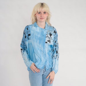 May include: A light blue button-up shirt with a tie-dye pattern and black floral print. The shirt has long sleeves and a relaxed fit.