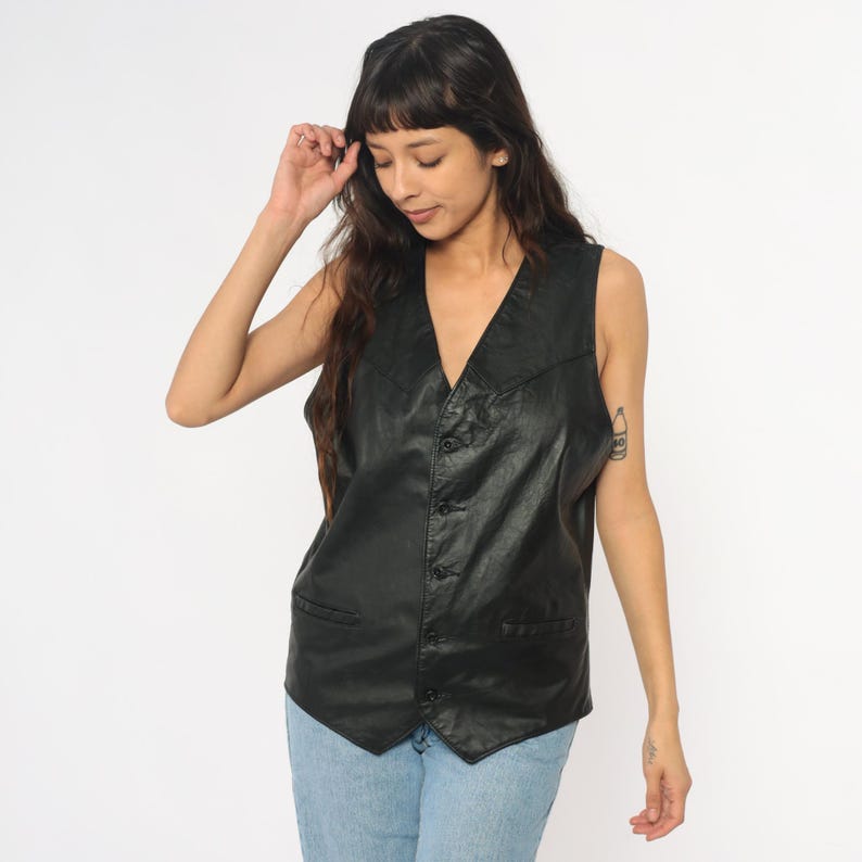 May include: A black leather vest with a V-neck and button closure. The vest has two front pockets and a tailored fit. The vest is sleeveless and appears to be made of a smooth, matte leather material. The vest is worn over a light blue denim jeans.