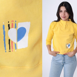 May include: Yellow sweatshirt featuring a graphic of a heart with a blue and white design. The heart is surrounded by vertical lines in black, pink and red.
