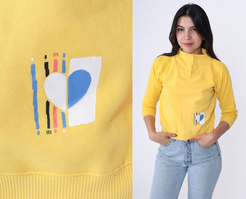 May include: Yellow sweatshirt featuring a graphic of a heart with a blue and white design. The heart is surrounded by vertical lines in black, pink and red.