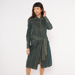 May include: A long denim dress in a medium wash, featuring a button-up front, long sleeves, and a matching belt. The dress has two front pockets and a knee-length hemline. The dress is made of a sturdy denim material.
