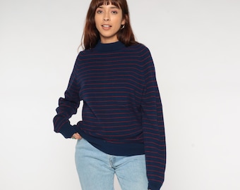 May include: Navy blue sweater with horizontal red stripes. The sweater has a mock neck and long sleeves. The model is wearing light blue jeans.