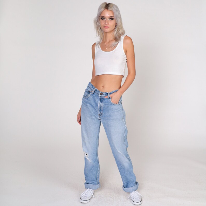 May include: A woman wearing a white vest top and light blue denim jeans with a distressed look. The jeans have a high waist and a relaxed fit.