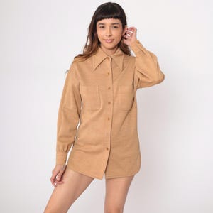 Puede incluir: Una camisa abotonada de color beige con mangas largas y dos bolsillos delanteros. La camisa tiene un cuello puntiagudo y está hecha de una tela texturizada. La prenda se usa como top y se combina con pantalones cortos.