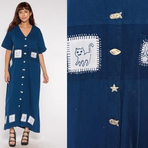 Op de afbeelding: Een donkerblauwe, knielange jurk met knopen, korte mouwen en een V-hals. De jurk heeft witte patches met katten- en visontwerpen en decoratieve knopen. De jurk wordt door een persoon gedragen.