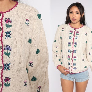 May include: A cream-colored knitted cardigan with a floral embroidered design. The cardigan has a button-up front and a pink crocheted trim around the neckline.