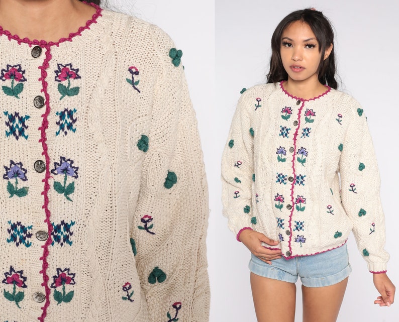 May include: A cream-colored knitted cardigan with a floral embroidered design. The cardigan has a button-up front and a pink crocheted trim around the neckline.