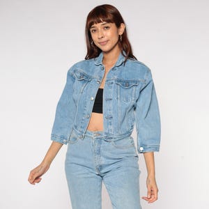 May include: Light blue denim jacket with three-quarter sleeves and a classic collar. The jacket has button closures and two chest pockets. The model is wearing a black top and light blue jeans.