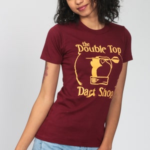 May include: A maroon t-shirt with a gold graphic of a hand throwing a dart at a dartboard. The graphic says "The Double Top Dart Shop".