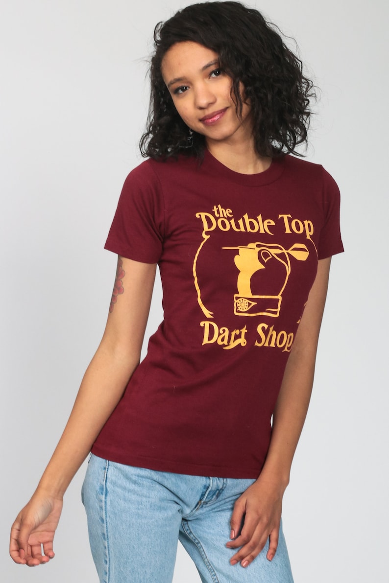 May include: A maroon t-shirt with a gold graphic of a hand throwing a dart at a dartboard. The graphic says "The Double Top Dart Shop".