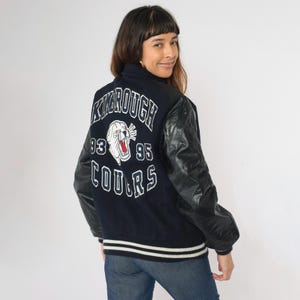 May include: A navy blue varsity jacket with black leather sleeves. The back of the jacket has a white and red embroidered logo that says "Kimbrough Cougars" with the years "93" and "95" on either side of the logo. The jacket has a white and black striped trim around the bottom and cuffs.