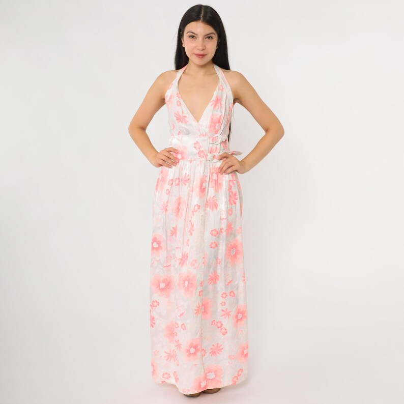 May include: A full-length, halter-neck dress with a white background and a pink floral pattern. The dress features a plunging neckline and a fitted waist with buckle details. The fabric appears to have a slight sheen, and the dress is floor-length.