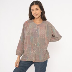 May include: A button-down shirt with a patchwork design in shades of purple, red, and green. The long-sleeved shirt has a round neckline and is worn with blue jeans. The shirt has a relaxed fit and a vintage aesthetic.