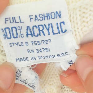 May include: Close-up of a white label with blue text. The label reads "FULL FASHION 100% ACRYLIC STYLE G 755/727 RN 34751 MADE IN TAIWAN R.O.C." The label is attached to a cream-colored knitted fabric.