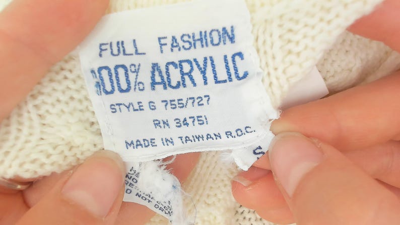 May include: Close-up of a white label with blue text. The label reads "FULL FASHION 100% ACRYLIC STYLE G 755/727 RN 34751 MADE IN TAIWAN R.O.C." The label is attached to a cream-colored knitted fabric.