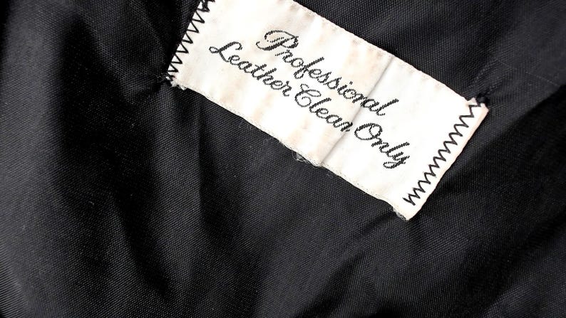 May include: Close-up of a black fabric with a white tag stitched to it. The tag reads "Professional Leather Clean Only" in black script. The tag has a zig-zag stitched border.