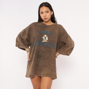 May include: An oversized brown acid-wash t-shirt with a crew neckline and short sleeves. The front features a graphic of a person sitting in a chair with blue text above and below the illustration that reads "KEEP EVERYTHING OK" and "I COULD SIT AND WATCH IT ALL DAY LONG".