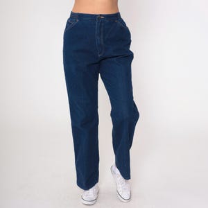 K&ouml;nnte beinhalten: Dunkelblaue Jeans mit hoher Taille und geradem Bein. Die Jeans hat einen Knopf- und Rei&szlig;verschluss sowie Taschen. Die Person tr&auml;gt wei&szlig;e Turnschuhe. Der Hintergrund ist schlicht wei&szlig;.