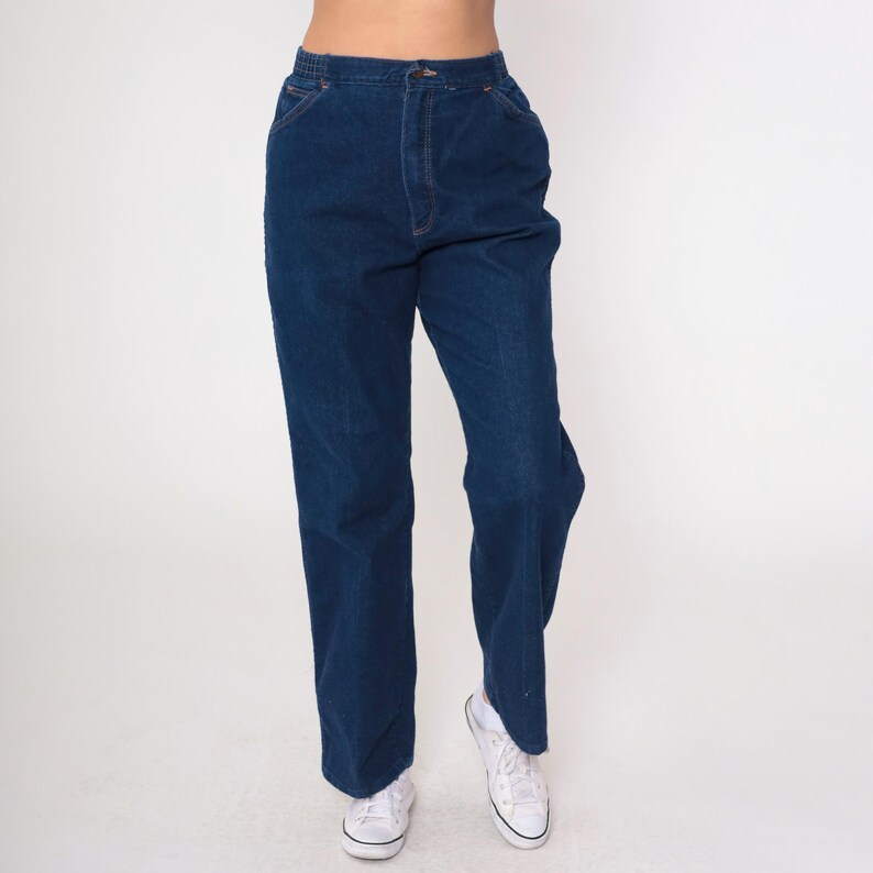 K&ouml;nnte beinhalten: Dunkelblaue Jeans mit hoher Taille und geradem Bein. Die Jeans hat einen Knopf- und Rei&szlig;verschluss sowie Taschen. Die Person tr&auml;gt wei&szlig;e Turnschuhe. Der Hintergrund ist schlicht wei&szlig;.
